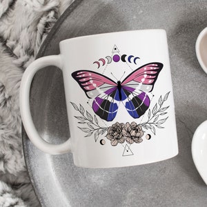 Może przedstawiać: Biały kubek ceramiczny z wzorem różowego, niebieskiego i czarnego motyla. Motyl jest otoczony geometrycznym wzorem z księżycami i kwiatami.