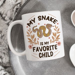 May include: White ceramic coffee mug with a brown snake illustration and the text "My Snake is my Favorite Child" in brown lettering with floral accents.