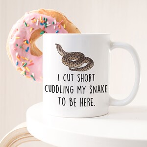 May include: White ceramic mug with a black snake illustration and the text "I cut short cuddling my snake to be here."