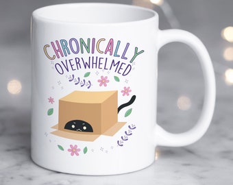 May include: White ceramic mug with the words "Chronically Overwhelmed" in colorful lettering. A cartoon cat peeks out from a cardboard box, surrounded by floral and leaf illustrations. The mug has a curved handle.