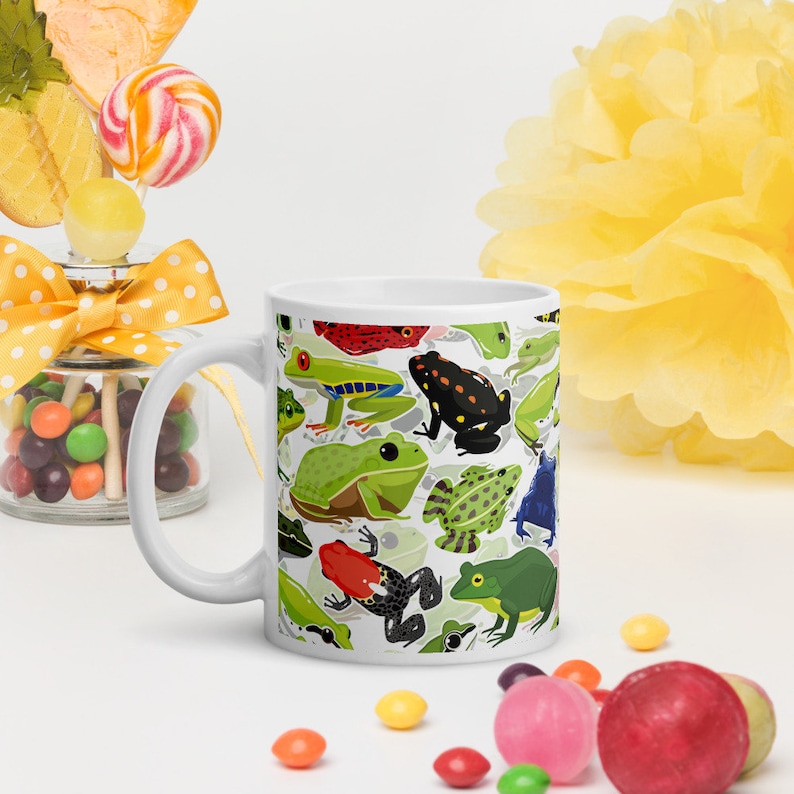 May include: White ceramic mug with a colorful pattern of frogs. The frogs are green, red, blue, black, and yellow. The mug is a standard size and has a handle.