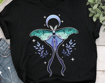 May include: Black t-shirt featuring a celestial moth design. The moth has teal and blue wings with a crescent moon and stars above. The design includes sprigs of lavender-colored leaves and additional stars.