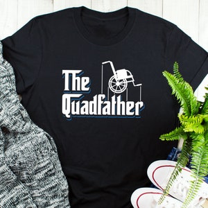 Puede incluir: Camiseta negra con un gráfico de silla de ruedas blanca y el texto "The Quadfather" en una fuente estilizada.