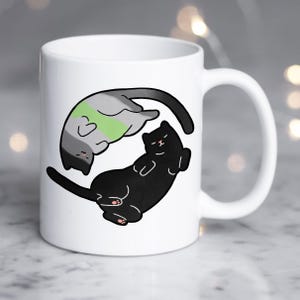 Pode incluir: Caneca de cerâmica branca com um gato preto e um gato cinza com uma faixa verde, dispostos em um design yin-yang. Os gatos têm olhos fechados e patas cor-de-rosa. A caneca tem uma alça branca.