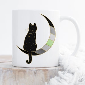 Puede incluir: Taza de cerámica blanca con un gato negro sentado en una luna creciente. La luna está decorada con un patrón de rayas verdes, grises y blancas.