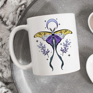 May include: White ceramic mug featuring a detailed illustration of a moth with purple, yellow, and black wings, a crescent moon, and stars. The moth design is centered on the mug, with delicate sprigs on either side. The mug has a curved handle.