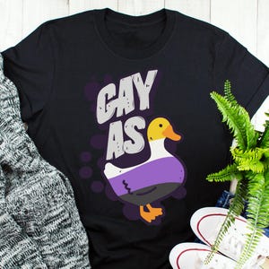 May include: Black t-shirt with the text "gay as" in white and a cartoon duck with purple, white, and black stripes.