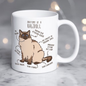 Pode incluir: Caneca de cerâmica branca com uma ilustração de gato Ragdoll e etiquetas anatômicas. O gato é marrom e creme, com olhos azuis. O texto inclui "Anatomia de um Ragdoll" e etiquetas humorísticas.