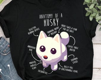 Puede incluir: Camiseta negra con una ilustración de dibujos animados de un perro husky y el texto "Anatomía de un Husky". El diseño incluye etiquetas que señalan varias características, como "Dispositivo de audición selectiva" y "Palos de zoom".