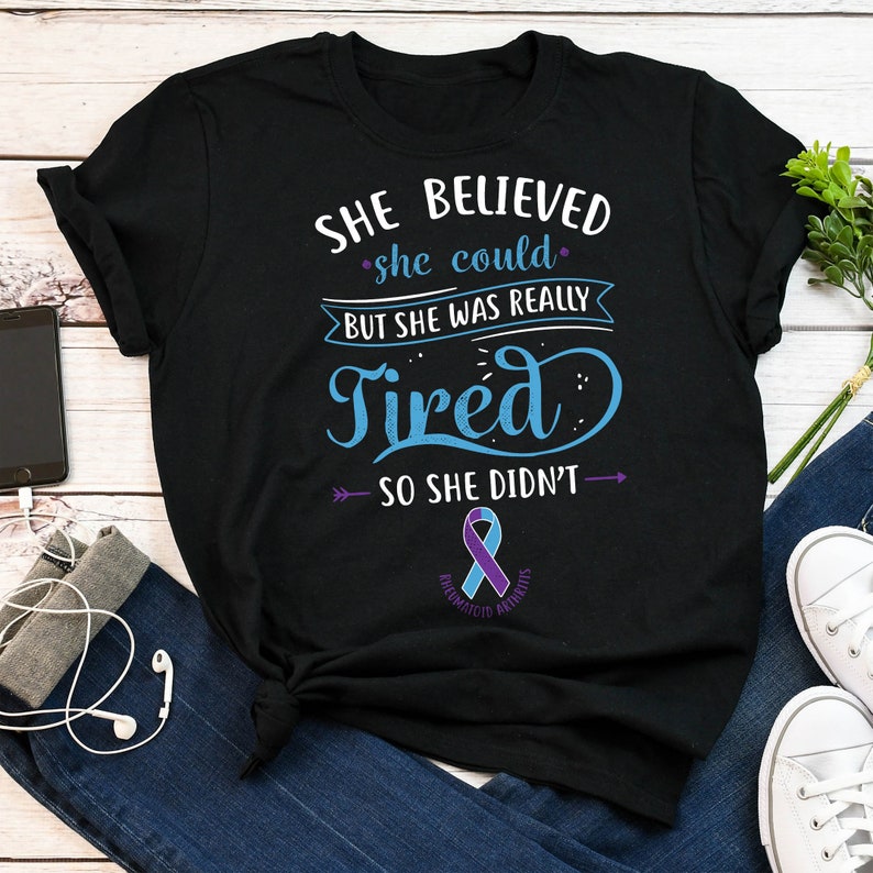 May include: Black t-shirt with a blue and purple ribbon and the text "SHE BELIEVED she could BUT SHE WAS REALLY Tired SO SHE DIDN'T" with arrows pointing to the left and right of the text. The ribbon has the words "RHEUMATOID ARTHRITIS" written on it.
