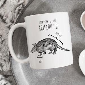 May include: White ceramic mug with a black and white illustration of an armadillo. The mug has the text "Anatomy of an Armadillo" and labels the armadillo's body parts "Arm" and "Adillo".