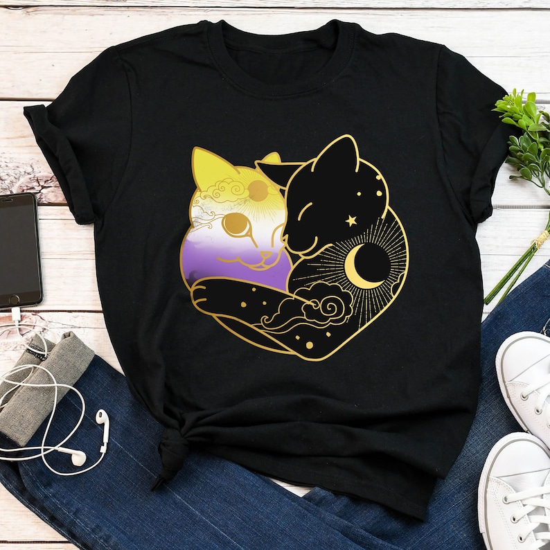 Op de afbeelding: Zwart T-shirt met een gouden omtrek van twee omhelzende katten. E&eacute;n kat is gekleurd met non-binaire pride vlag kleuren, de andere is zwart met een hemels ontwerp. Het ontwerp bevat een zon, maan, sterren en wolken.