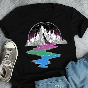 Op de afbeelding: Zwart T-shirt met een witte afbeelding van een bergketen met een rivier die erdoorheen stroomt. De rivier is een gradiënt van blauw, groen en paars. De bergketen is omringd door een cirkel van sterren.