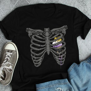 May include: Black t-shirt featuring a graphic of a ribcage with a heart inside. The heart is a gender-neutral pride flag with yellow, white, purple, and black stripes.