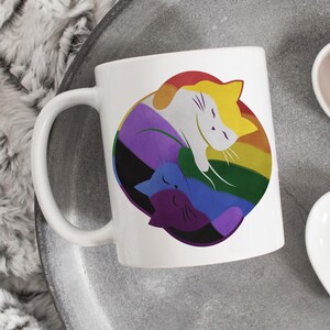 Puede incluir: Taza de cerámica blanca con una colorida ilustración de dos gatos estilizados dentro de un círculo arcoíris. Los gatos son en tonos de amarillo, morado, azul y blanco. La taza tiene un asa curva y está sobre una superficie gris.