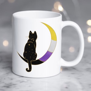 Könnte beinhalten: Weiße Keramiktasse mit einer schwarzen Katzensilhouette, die auf einem Halbmond sitzt. Der Mond ist mit den Farben der Non-Binary-Pride-Flagge verziert: Gelb, Weiß, Lila.