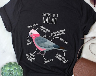 May include: Black t-shirt featuring a detailed illustration of a Galah parrot with labeled anatomical features. The design includes text such as "Anatomy of a Galah," "Chaos-ometer," and "Magical Cotton Candy Plumage."