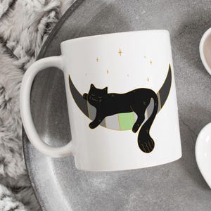 Pode incluir: Caneca de cerâmica branca com um gato preto dormindo em uma lua crescente com estrelas douradas. O gato tem uma faixa verde nas costas.