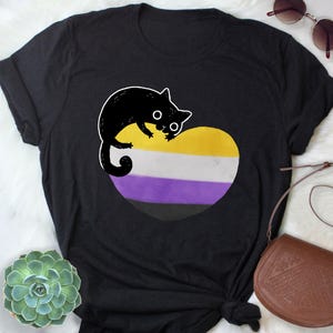 May include: Black t-shirt featuring a cartoon black cat perched on a heart-shaped design. The heart displays the non-binary pride flag colors: yellow, white, purple, and black. The cat has large, round eyes and a playful expression.