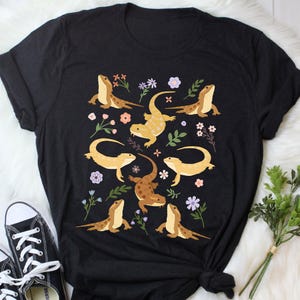 Puede incluir: Camiseta negra con un patrón colorido de dragones barbudos y flores. Los dragones barbudos están en varias poses, algunos miran hacia adelante, otros miran hacia los lados y otros miran hacia atrás. Las flores son de varios colores, incluyendo rosa, morado y amarillo.