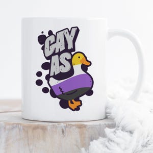 May include: White ceramic mug with a black and white illustration of a duck with a purple, white, and yellow stripe pattern. The text "gay as" is written in black and white above the duck.