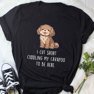 Puede incluir: Camiseta negra con una imagen de dibujos animados de un perro cavapoo marrón y blanco. El texto en la camiseta dice "I cut short cuddling my cavapoo to be here."