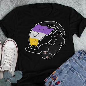 Pode incluir: Camiseta preta com um gráfico de dois gatos, um preto e outro com listras roxas, amarelas e brancas. Os gatos estão enroladas um ao redor do outro em um símbolo yin-yang.