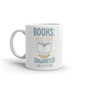 Puede incluir: Taza de cer&aacute;mica blanca con el texto "BOOKS: YOUR BEST DEFENSE AGAINST UNWANTED CONVERSATION" en azul y negro. El dise&ntilde;o incluye una ilustraci&oacute;n de un libro abierto con l&iacute;neas radiantes.