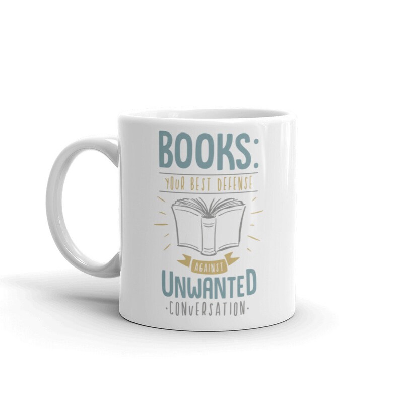 Puede incluir: Taza de cer&aacute;mica blanca con el texto "BOOKS: YOUR BEST DEFENSE AGAINST UNWANTED CONVERSATION" en azul y negro. El dise&ntilde;o incluye una ilustraci&oacute;n de un libro abierto con l&iacute;neas radiantes.