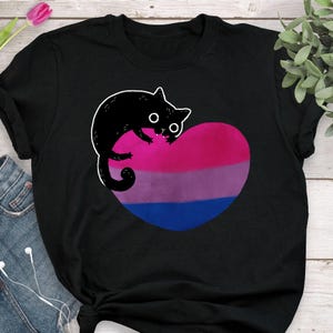 以下が含まれることがあります： 黒いTシャツ。漫画の猫のデザインが特徴です。 白いアウトラインの黒い猫が、バイセクシュアルプライドフラッグの色（ピンク、紫、青）のハート型のグラフィックの上にいます。 シャツは柔らかい素材で作られています。