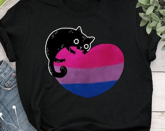May include: Black t-shirt featuring a cartoon cat design. The cat is black with white outlines and is perched on a heart-shaped graphic in the colors of the bisexual pride flag: pink, purple, and blue. The shirt is made of a soft material.