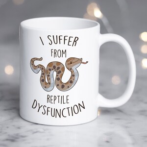 May include: White ceramic mug with a black and white illustration of a snake and the text "I suffer from reptile dysfunction".