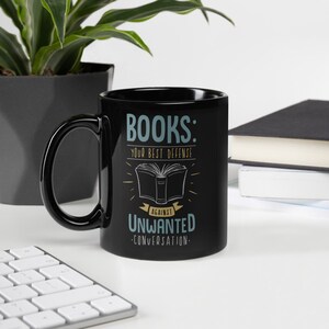Puede incluir: Taza de cer&aacute;mica negra con el texto "BOOKS: YOUR BEST DEFENSE AGAINST UNWANTED CONVERSATION" en blanco y turquesa. El dise&ntilde;o incluye una ilustraci&oacute;n de un libro abierto. La taza est&aacute; sobre un escritorio blanco con un teclado y una planta de fondo.