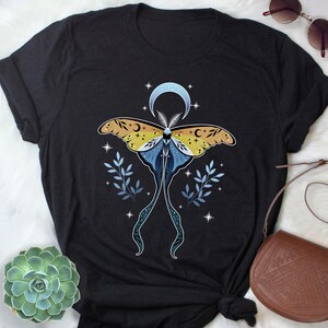 May include: Black t-shirt with a celestial moth design. The moth has yellow and blue wings with crescent moon and star details. The design includes a crescent moon, stars, and leafy sprigs.