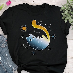 May include: Black t-shirt with a graphic of a cat floating in space with a galaxy design. The cat's tail is yellow and the background is filled with stars and planets.
