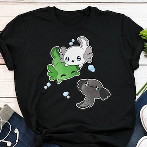 Può includere: Maglietta nera con una grafica di tre axolotl. Gli axolotl sono verdi, bianchi e neri. Gli axolotl sono in stile cartone animato e hanno occhi grandi.