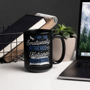 May include: Black coffee mug with a white design that says "I am too emotionally attached to fictional characters". The mug has a white star and a small horse graphic on the design. The mug is sitting on a white surface.