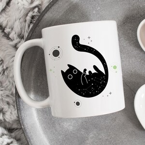 Pode incluir: Caneca de cerâmica branca com uma silhueta preta de um gato com estrelas dentro de seu corpo. O gato está enroscado em uma bola com sua cauda enroscada ao redor de seu corpo. O fundo é branco com pequenas estrelas pretas e verdes.