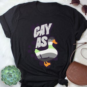 May include: Black t-shirt with the text "gay as" in white and a cartoon duck with a green, gray, and black pattern.