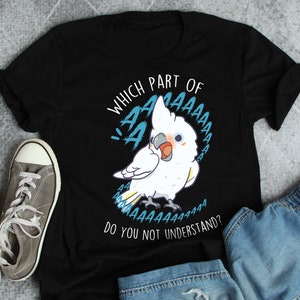 May include: Black t-shirt featuring a cartoon white cockatoo with blue accents and the text "WHICH PART OF AAAAAAAA DO YOU NOT UNDERSTAND?" The bird has a blush on its cheeks. A pair of gray sneakers and denim shorts are in the background.
