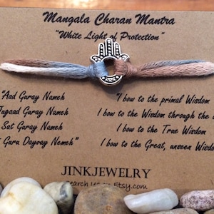 May include: A brown card with the text "Mangala Charan Mantra" and "White Light of Protection" printed on it. A silver Hamsa hand charm is attached to a braided cord with three colours: brown, white, and blue. The card also includes the text "Aad Guray Namek, Jugaad Guray Nameh, Sat Guray Nameh, Siri Gara Dayvay Nemek" and "I bow to the primal Wisdom, I bow to the Wisdom through the ages, I bow to the True Wisdom, I bow to the Great, unseen Wisdom". The card also includes the text "JINKJEWELRY" and "Search Etsy.com".