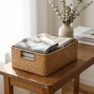 Puede incluir: Una cesta rectangular tejida con ropa de cama gris y beige doblada. La cesta tiene asas recortadas y está sobre una mesa de madera. Un jarrón con vegetación y libros están en el fondo.