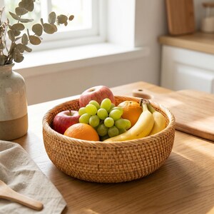 May include: A round, woven wicker fruit basket filled with fresh fruit. The basket contains red apples, green grapes, oranges, and bananas. The basket is sitting on a light-colored wooden surface, with a vase and window in the background.