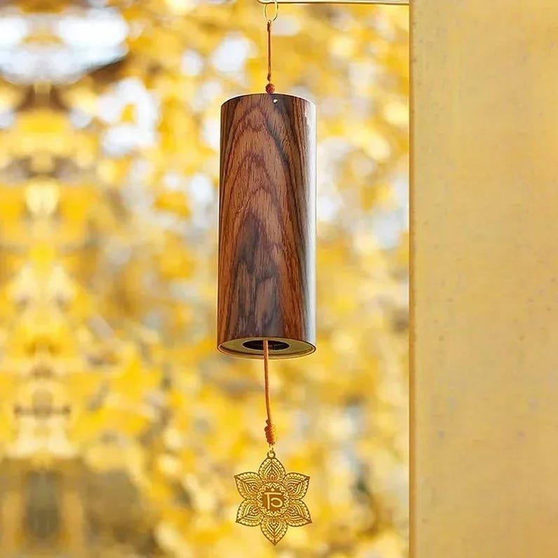 May include: A cylindrical wind chime made of dark wood with a gold-colored floral pendant. The wind chime hangs from a thin metal hook. The wood grain is visible on the surface of the chime. The pendant has a detailed, openwork design.