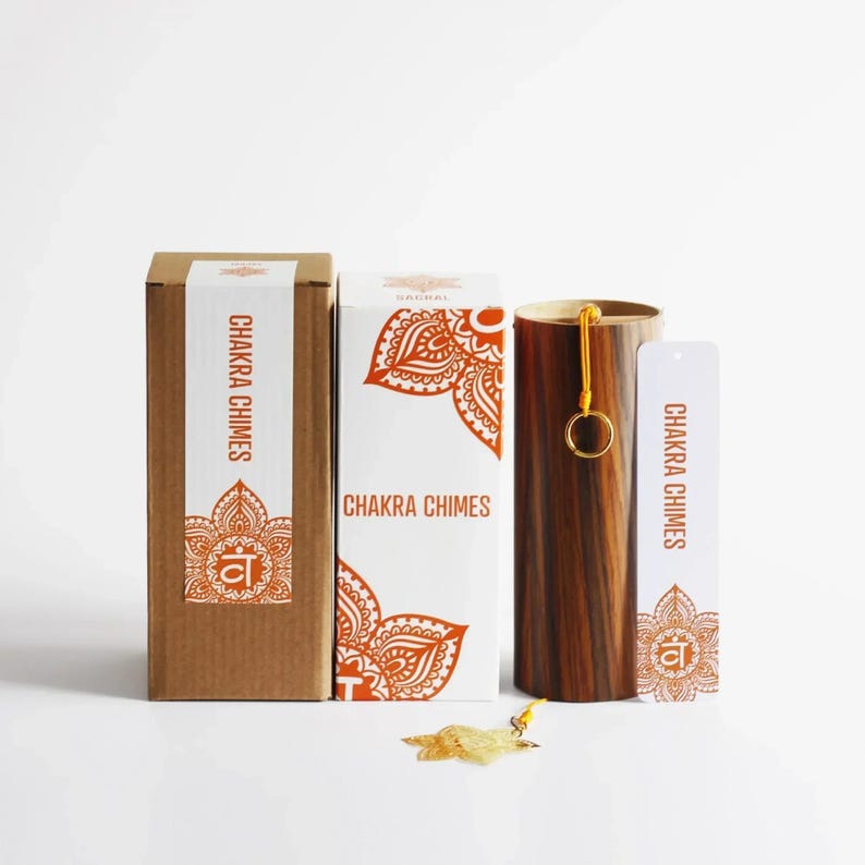 May include: A set of Chakra Chimes, featuring a wooden chime, two boxes, and a tag. The boxes are white and brown with orange mandala designs and the text "Chakra Chimes". The wooden chime is cylindrical with a gold ring and yellow cord.