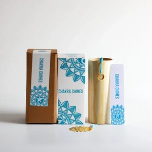 May include: A set of Chakra Chimes, including a cylindrical wooden chime, two boxes, and a tag. The boxes and tag feature a teal mandala design and the words "Chakra Chimes". A gold-colored decorative element is at the base.