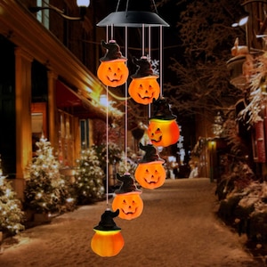 May include: A Halloween-themed wind chime featuring six orange jack-o'-lanterns wearing black witch hats. The smiling pumpkins are suspended from a black solar panel, creating a festive outdoor decoration. The background shows a street scene.