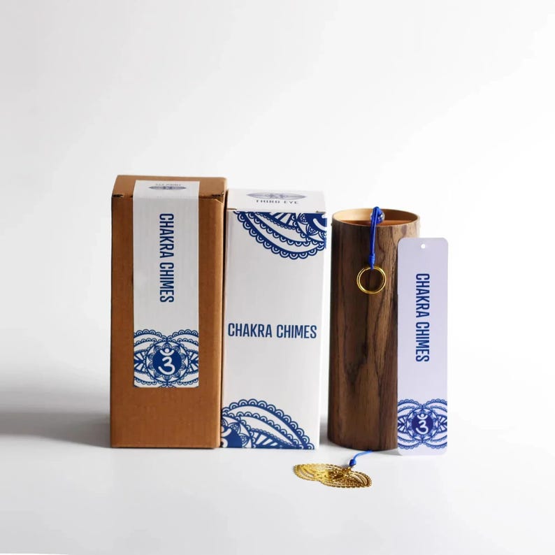 May include: A set of chakra chimes with packaging. The chimes are in a wooden cylinder with a blue cord and gold ring. The packaging includes two boxes and a bookmark, all with the text "Chakra Chimes" and blue decorative patterns.