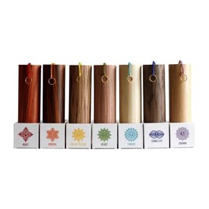 May include: A set of seven wooden chakra boxes with varying wood grains and colors, each with a gold ring and colored cord. Each box sits on a white base with a chakra symbol and name printed on it.