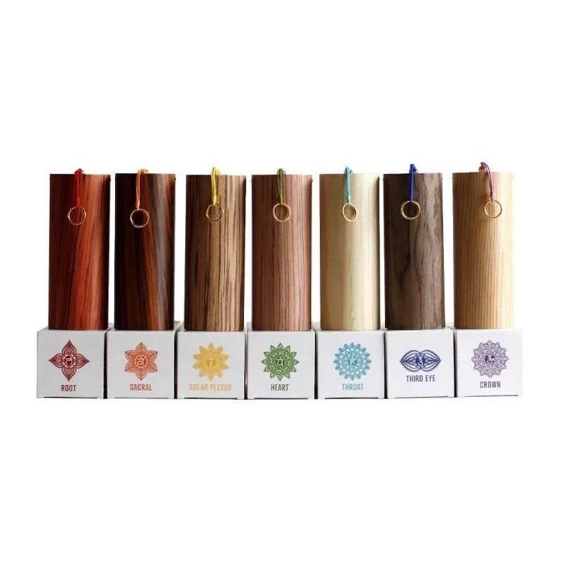 May include: A set of seven wooden chakra boxes with varying wood grains and colors, each with a gold ring and colored cord. Each box sits on a white base with a chakra symbol and name printed on it.